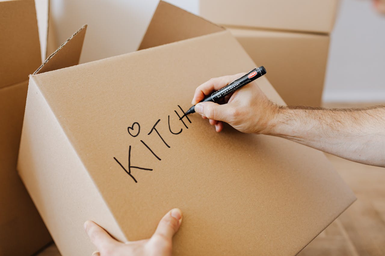 From above of unrecognizable adult male writing with black marker on cardboard containers while packing staff and preparing for relocation in new home